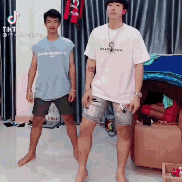 two men are standing next to each other in a living room . one is wearing a balenciaga shirt .