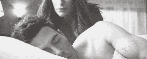a black and white photo of a woman standing next to a man sleeping on a bed .