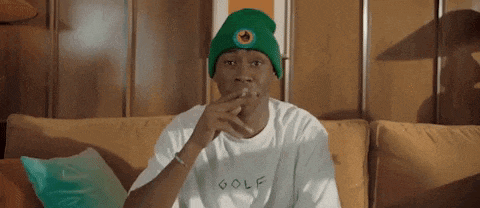 a man wearing a green beanie and a white t-shirt is sitting on a couch smoking a cigarette .