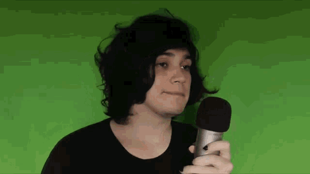 a young man with long black hair is holding a microphone in front of a green screen .