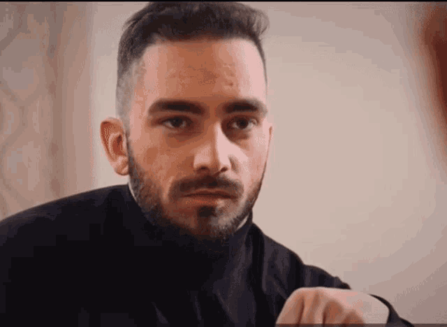 a man with a beard wearing a black turtleneck is looking at the camera