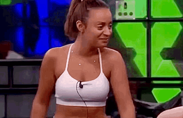 a woman in a white sports bra with a microphone on her neck is sitting in front of a green wall .