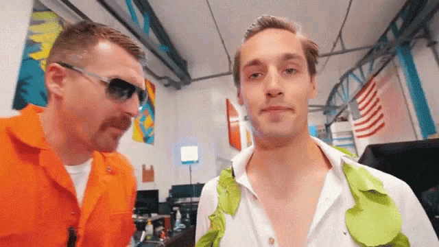 two men are standing next to each other in a room . one of the men is wearing sunglasses and a white shirt .