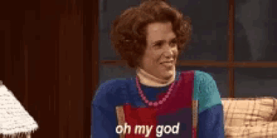 a woman is sitting on a couch wearing a blue sweater and a red apron and says `` oh my god '' .
