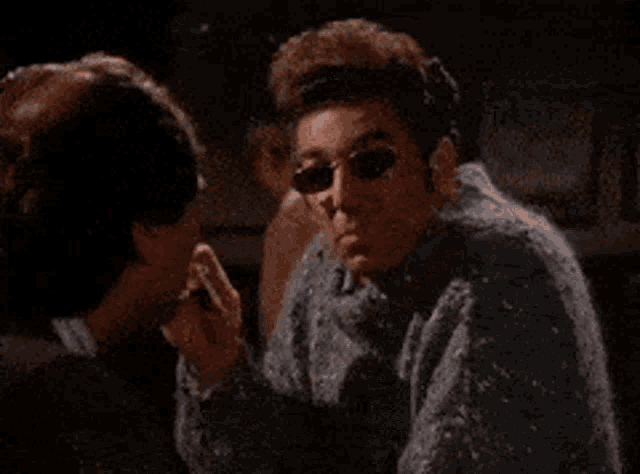 a man wearing sunglasses is smoking a cigarette while another man drinks from a glass .