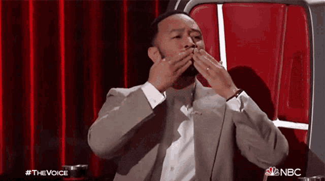 a man in a suit is covering his mouth with his hands while sitting in front of a red curtain on nbc
