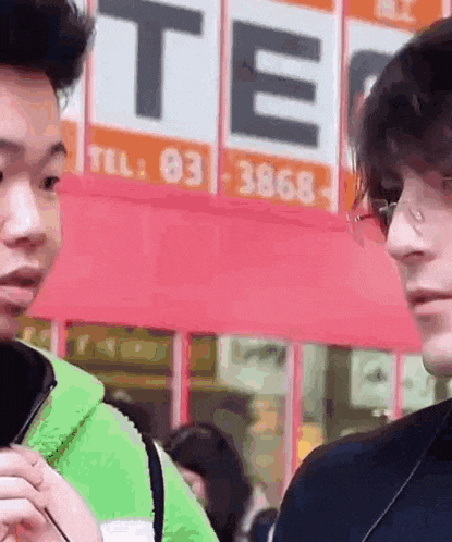 a man wearing glasses is talking to another man in front of a sign that says tel . 03 3868 .