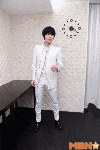 a man in a white suit stands in front of a wall clock with mbn written on it