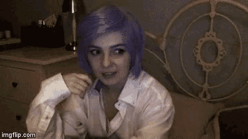 a woman with purple hair and a white shirt is sitting on a bed and looking at the camera .