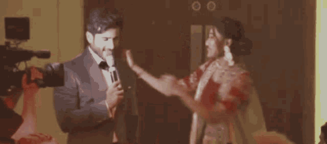 a man in a tuxedo is talking into a microphone while a woman in a white dress looks on