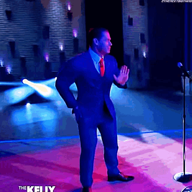 a man in a suit and tie is standing in front of a microphone with the kelly logo on the bottom