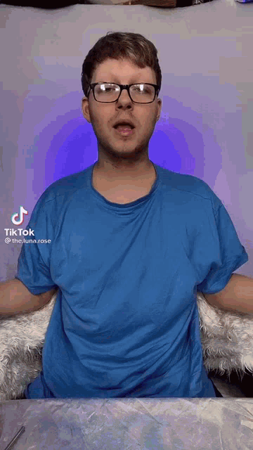 a man wearing glasses and a blue shirt is sitting at a table with his hands outstretched .