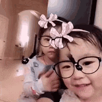 a little girl wearing glasses and a headband with a pink bow is taking a selfie with another little girl .