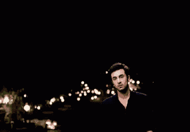 a man in a black shirt is standing in the dark in front of a blurry background of lights .