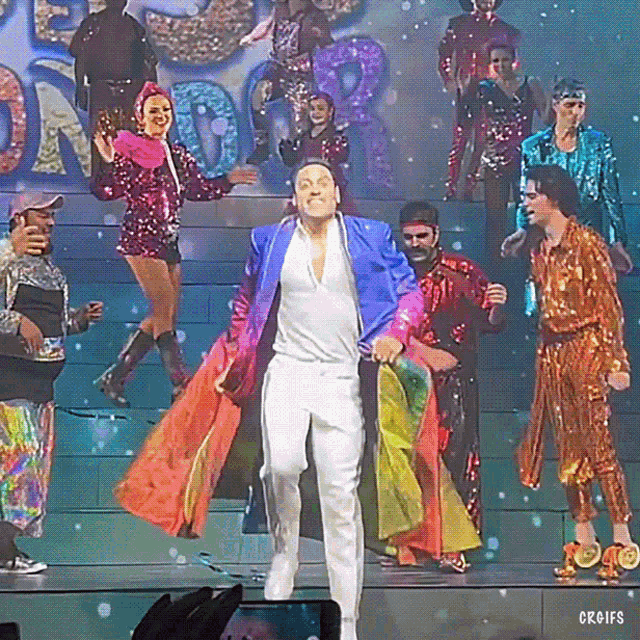 a man in a blue jacket stands on a stage surrounded by sequined people