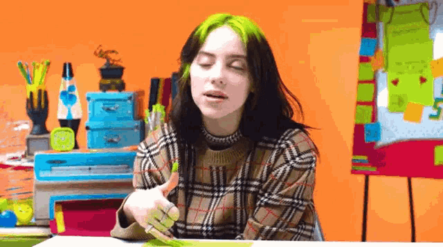 a woman with green hair is sitting at a desk with her eyes closed and holding a marker .
