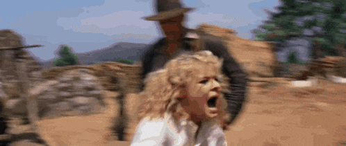 a woman is screaming while a man in a hat stands behind her in the desert .