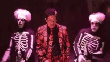a man in a red suit is standing between two skeletons on a stage .