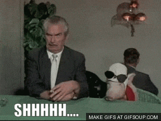 a man in a suit and tie is smoking a cigarette while a stuffed animal wearing sunglasses looks on behind him