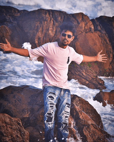 a man in a pink shirt and ripped jeans is standing on a rock near the ocean