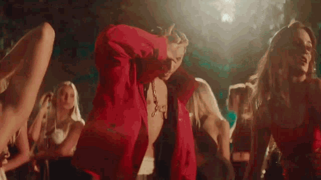 a shirtless man in a red jacket is dancing with a group of women at a party .