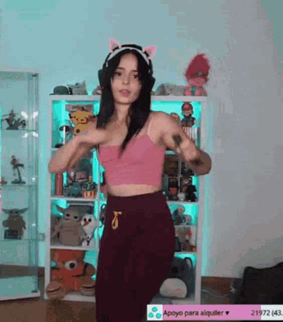 a woman in a pink top and burgundy pants is dancing in front of a shelf with stuffed animals on it