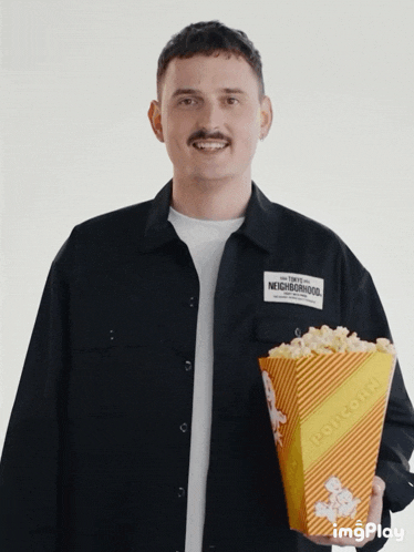 a man wearing a black neighborhood shirt is holding a yellow striped container of popcorn