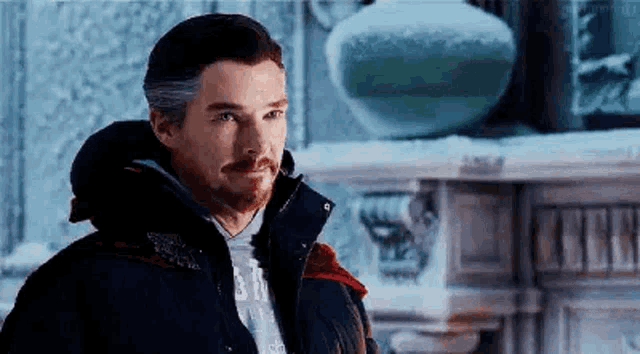 a man with a beard is wearing a jacket and standing in front of a fireplace in the snow .