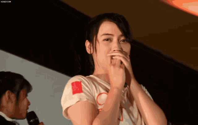 a woman covering her mouth with her hands while wearing a white shirt with a red circle on it