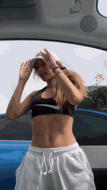 a woman in a crop top and white sweatpants is standing in the back of a blue car .