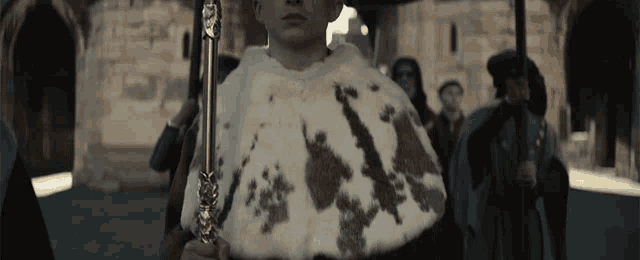 a man in a fur cape is holding a sword in front of a crowd of people .