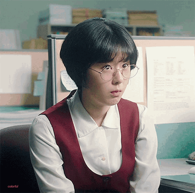 a woman wearing glasses and a red vest sits at a desk with colorful written on the bottom