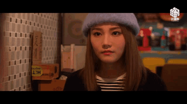 a woman wearing a purple hat looks at the camera in front of a sign that says hkt