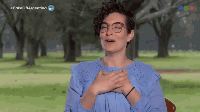 a woman wearing glasses and a blue shirt holds her hands to her chest in front of a park with the words bake off argentina above her