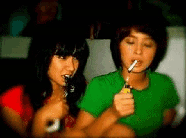 a woman in a green shirt is lighting a cigarette next to a woman in a red shirt .
