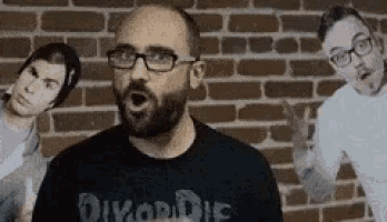 a man with glasses is standing in front of a brick wall with two other men .
