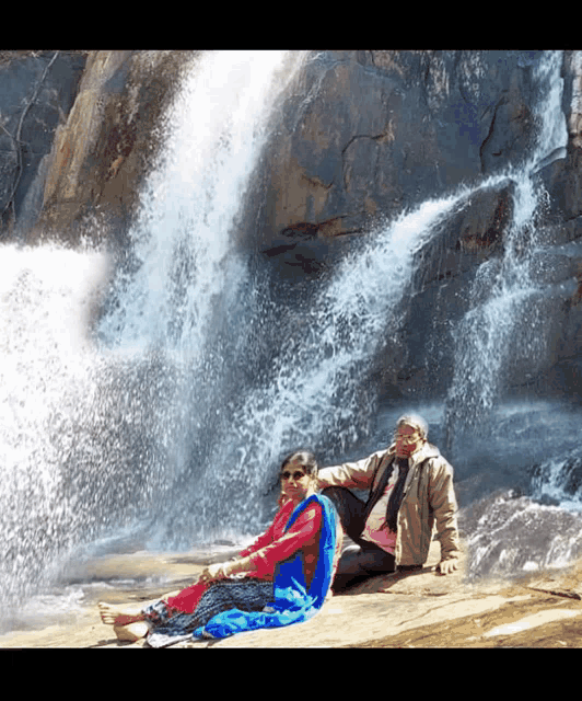 a man and a woman are sitting on a rock in front of a waterfall