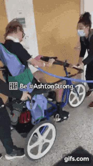 a person in a wheelchair is being pushed by a person wearing a mask and a sign that says be #ashtonstrong