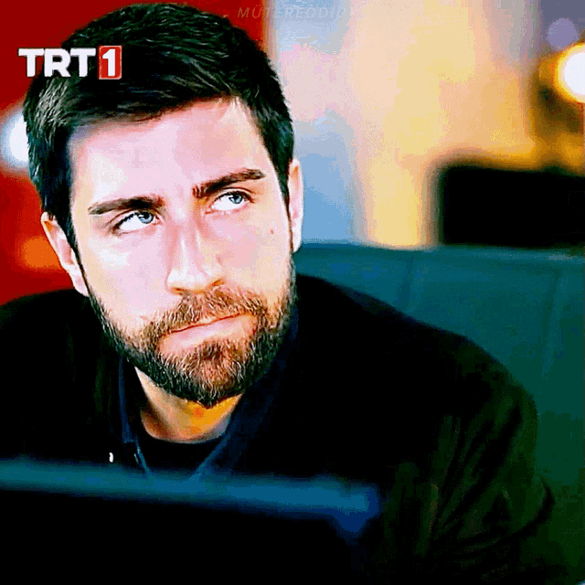 a man with a beard is sitting in front of a trt 1 screen