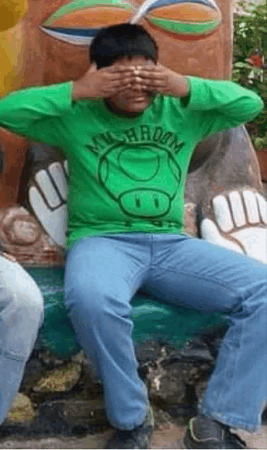 a person wearing a green shirt that says mushroom is covering his eyes