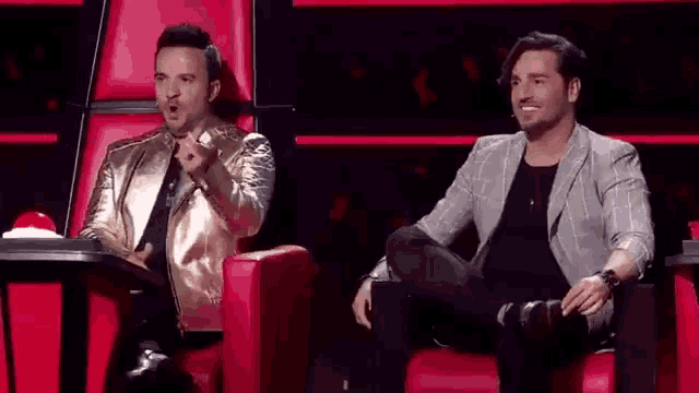 two men are sitting in red chairs on a stage and one of them is pointing at the camera .