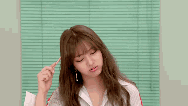 a woman holds a pen to her forehead in front of a green blind