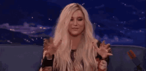 a woman with blonde hair is sitting on a couch with her hands up and the word demais written on the screen behind her .