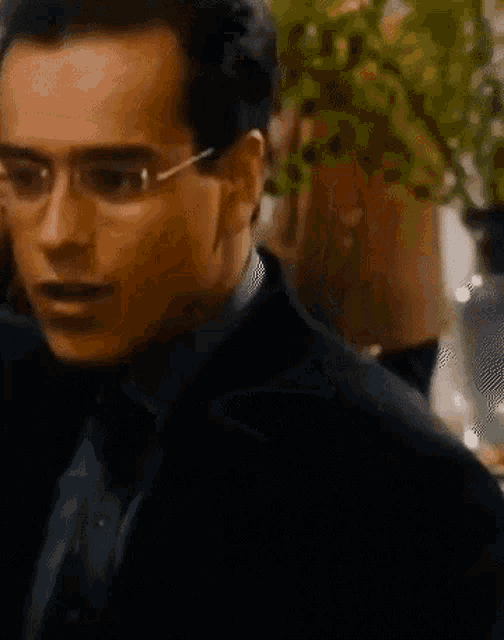 a man wearing glasses and a suit is standing in front of a plant .