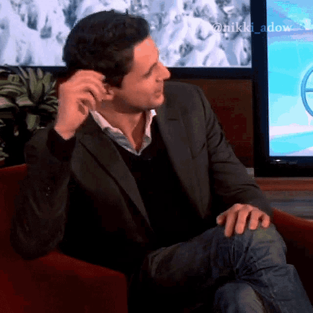 a man in a suit sits in front of a television with the name nikki adow on the bottom right