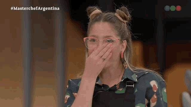 a woman wearing glasses is covering her mouth with her hand while watching a show called masterchef argentina