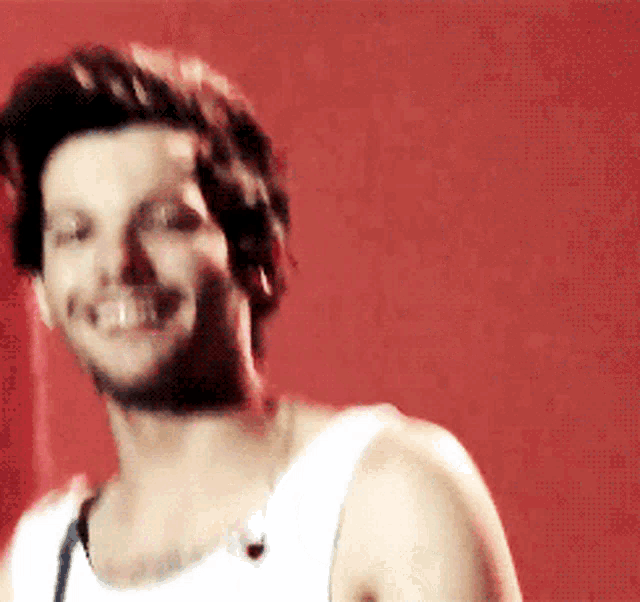 a man in a white tank top smiles in front of a red wall