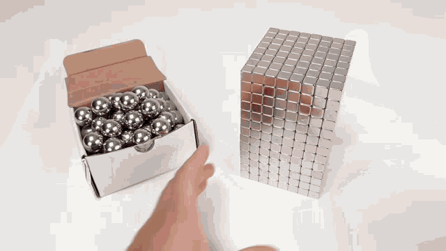 a box of metal balls sits next to a person holding a metal cube