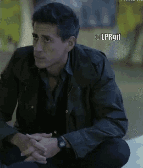 a man in a green jacket sits on the ground with his hands folded and a lprgif watermark above him
