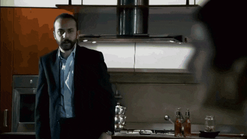 a man in a suit stands in a kitchen looking at another man